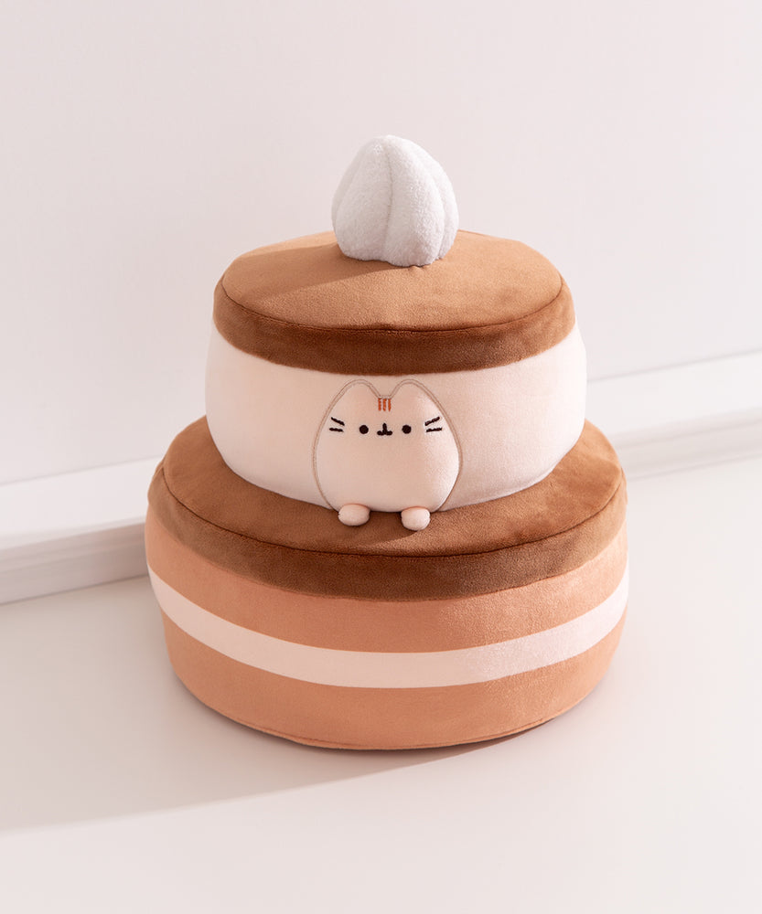 Pusheen Cake Plush is a layered chocolate cake with alternating light brown cake, white cream, and dark brown icing layers. The cake is topped with a whipped cream plush dollop.