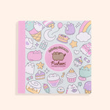 Front cover of the Coloring Cuteness: A Pusheen Coloring & Activity Book. The front cover includes graphics of food items and Pusheen the Cat in various forms including a cupcake, a ghost, a cinnamon roll, a pancake stack, a carton of strawberry milk, and more. In the center of the cover is a pink circle outlined in white that states the title of the book and the author, Claire Belton. The spine of the book is light pink and the background graphic color is mint green. 