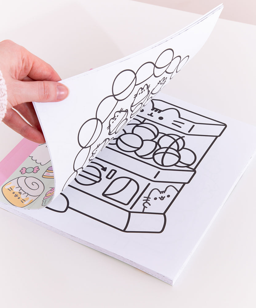 Model holds open the Pusheen activity book showcasing one of the coloring pages. The open page shows a black outline Pusheen as a vending machine with various spheres inside. The vending machine has a turn knob and opening slot for the user to grab the ball. The black outlined graphic is printed in the center of the white page.  