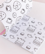 Pusheen Coloring & Activity Book is opened to two white coloring pages with black graphics. On the left page is a repeating pattern of Pusheen as a donut surrounded by cups of coffee and smaller glazed donuts. On the righthand page is a repeating pattern of Pusheen the Cat sitting atop a macaron surrounded by polka dots, tea pots, and smaller macarons.  