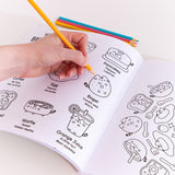Model holds a yellow-colored pencil above a graphic of various breakfast items on a page of the Pusheen coloring book. The book is open to show the lefthand page graphic and part of the righthand page. On the lefthand page showcases Pusheen as eight different breakfast items with character descriptors under each. On the righthand page is a circular graphic where Pusheen takes the form of an avocado toast, bagel, cereal, and sunny-side-up egg accompanied by forks, spoons, strawberries, and sparkles. 