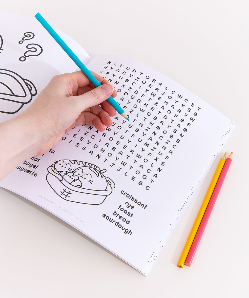 Model holds a blue colored pencil above a crossword puzzle page of the Pusheen activity book. The crossword puzzle has many bread-themed words including croissant. Sourdough, and toast. Under the crossword puzzle is a graphic of a breadbasket holding Pusheen taking the form of a baguette, croissant, and loaf of bread.  