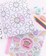 Interior page view of the Pusheen coloring book. A graphic of Pusheen lying in the center of a flower is partially colored using colored pencils that are laid out to the side of the coloring book. Below the interior page of the activity book is another copy of the book showing the pastel-colored front cover. Next to the books are paper clips, a pencil sharpener, an eraser, and three colored pencils. 