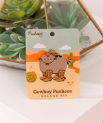 Large pusheen pin has gold foil outline that highlights the shape of the pin. The paper backer card that the pin is attached to has a pink and orange desert scene with green cacti and yellow sand.