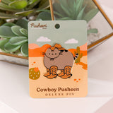 Large pusheen pin has gold foil outline that highlights the shape of the pin. The paper backer card that the pin is attached to has a pink and orange desert scene with green cacti and yellow sand.