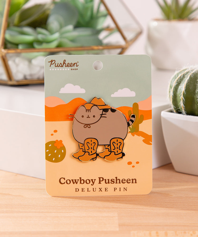 Front view of the Cowboy Pusheen Boots Deluxe Pin. Pusheen the cat wears a brown cowboy hat with four brown boots. The grey and brown cat pin has gold plating outlines and is held in the center of a desert scene backer card.