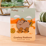 Front view of the Cowboy Pusheen Boots Deluxe Pin. Pusheen the cat wears a brown cowboy hat with four brown boots. The grey and brown cat pin has gold plating outlines and is held in the center of a desert scene backer card.