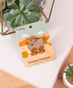 Cat shaped Pusheen pin lies on a white and brown surface surrounded by cactus plants.