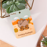 Cat shaped Pusheen pin lies on a white and brown surface surrounded by cactus plants.