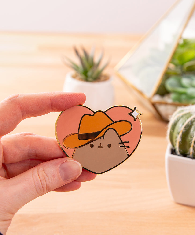 Model holds the Cowboy Pusheen Heart Deluxe Pin to show the scale of the heart-shaped pin with gold plated outline details.