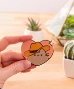 Pusheen the Cat wears a brown cowboy hat and smiles. Pusheen's classic whiskers and head stripes are present on this pin.