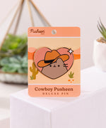 Front view of the Cowboy Pusheen Heart Deluxe Pin. Pusheen the cat wears a brown cowboy hat. The grey, brown, pink, and white pin has gold plating outlines and is held in the center of a desert scene backer card.