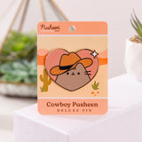 Front view of the Cowboy Pusheen Heart Deluxe Pin. Pusheen the cat wears a brown cowboy hat. The grey, brown, pink, and white pin has gold plating outlines and is held in the center of a desert scene backer card.