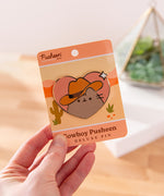 Model holds backer card of Pusheen Cowboy Pin. The paper backer card has a pink, orange, and yellow desert motif with green cactuses. 