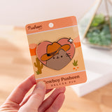 Model holds backer card of Pusheen Cowboy Pin. The paper backer card has a pink, orange, and yellow desert motif with green cactuses. 