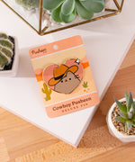 Heart shaped Pusheen pin lies on a white and brown surface surrounded by cactus plants.