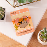 Heart shaped Pusheen pin lies on a white and brown surface surrounded by cactus plants.