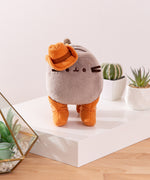 Front view of Cowboy Pusheen Plush. The grey fluffy Pusheen stands on all four paws in cowboy boots in front cacti. The cat plush is topped with a cowboy hat.