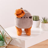 Front view of Cowboy Pusheen Plush. The grey fluffy Pusheen stands on all four paws in cowboy boots in front cacti. The cat plush is topped with a cowboy hat.