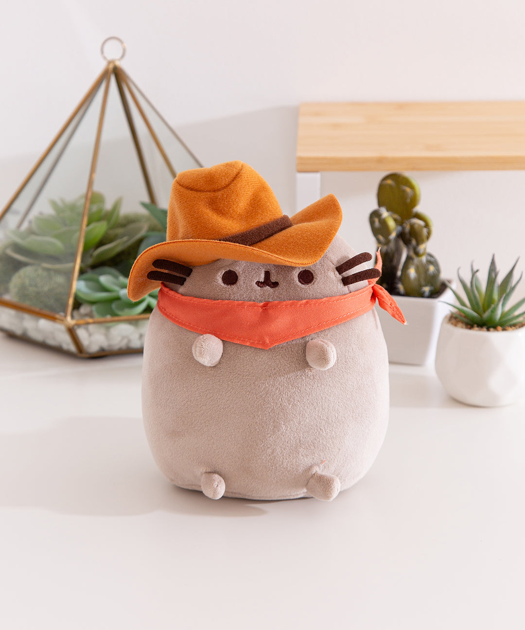 Front view of the Cowboy Pusheen Squisheen Plush. The classic Pusheen has dark brown embroidery for her mouth and eyes as well as dark brown fabric whiskers. The gray tabby cat wears and orange bandana around her neck and a brown hat atop her head.