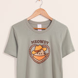 Green t-shirt with a large graphic on the front center. The graphic shows Pusheen the Cat wearing a brown cowboy hat and orange bandana in a heart-shaped rope.