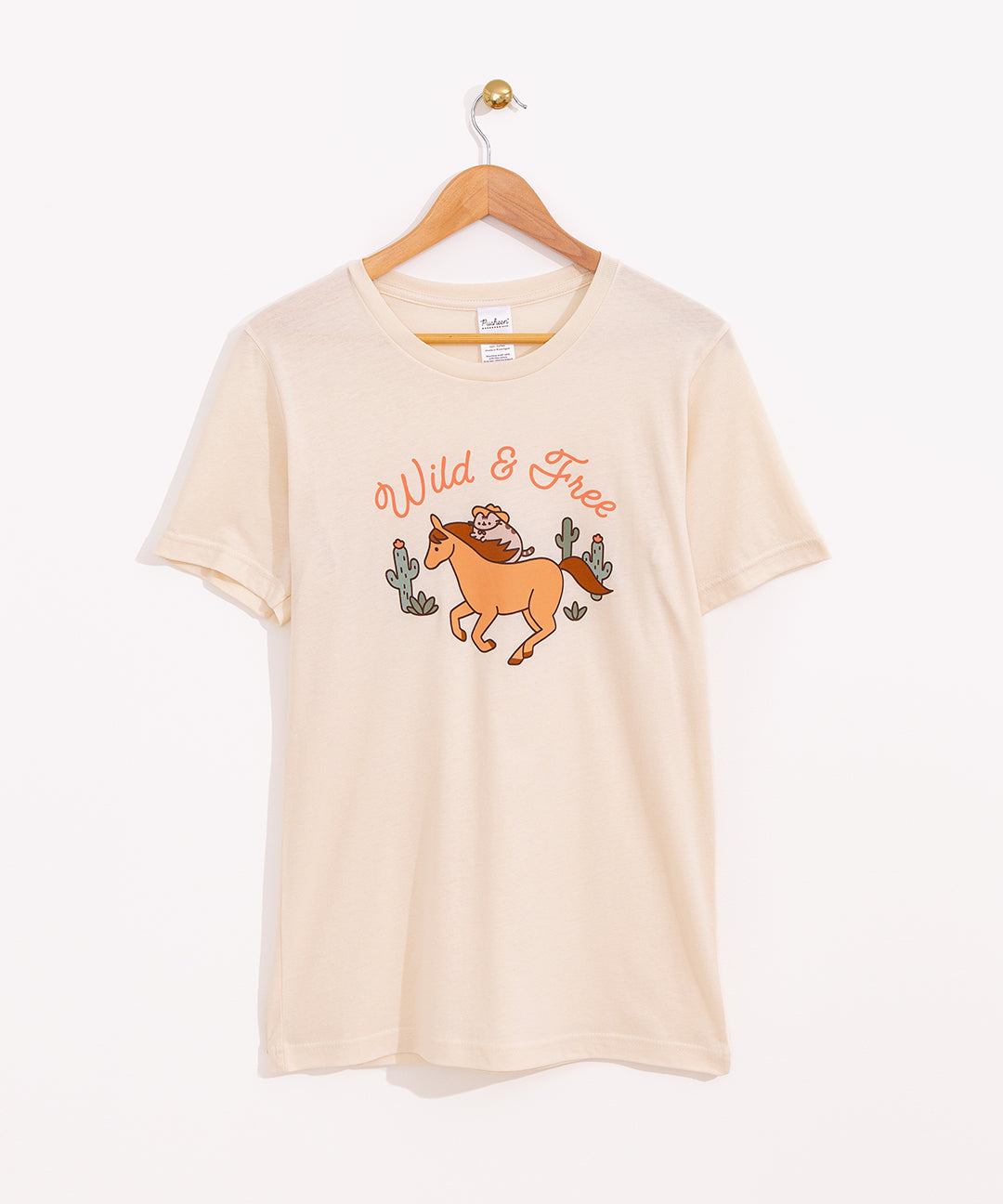 Cowboy Pusheen Wild & Free Tee hangs on a light brown hanger on a light background. The beige tee shirt has a graphic of Pusheen the Cat riding a brown horse in the desert.