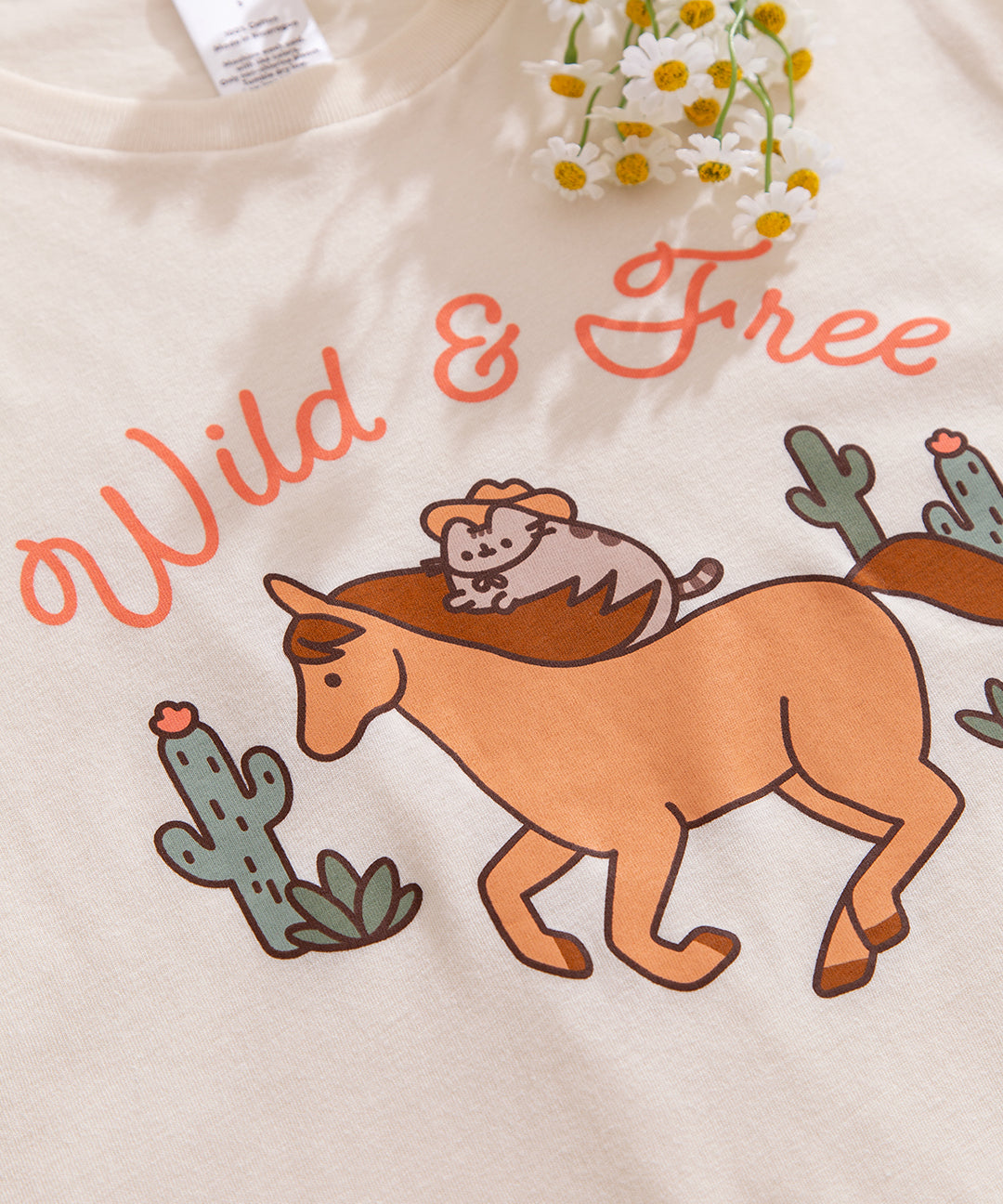 Close-up view of cowboy Pusheen graphic tee. The phrases above Pusheen desert graphic in orange colored font is "Wild & Free"