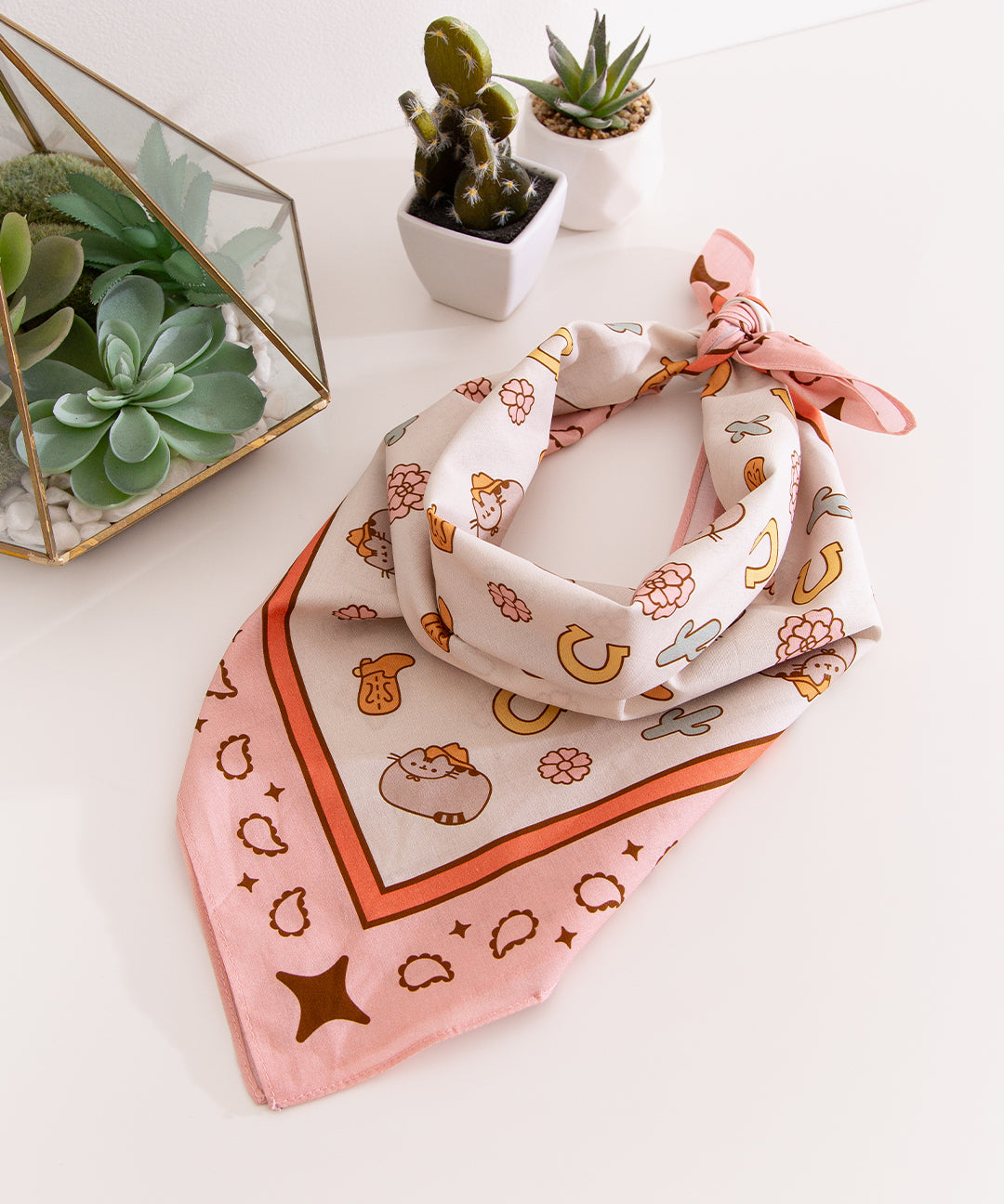 Cowboy Pusheen Bandana folded as a neck scarf. The light beige scarf with pink trim has graphics of flowers, paisley, catci, cowboy boots, horseshoes, and pusheen the cat wearing a cowboy hat.