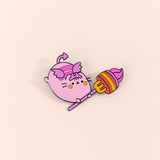 Front view of the Devil’s Food Cake Pusheen Deluxe Pin. Pin features a light pink Pusheen with purple wings, a purple forked tail, a purple crown, and a purple and yellow fork with a devil’s food cake pierced. The devil's food cake is shown in brown, yellow, pink, and purple layers. The pin sits in front of a yellow background. 
