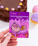 Model holds Devil’s Food Cake Pusheen Pin on its purple-colored backer card that features many layered cakes. In the background sits a chocolate cake and vanilla cupcakes with purple and pink frosting, 