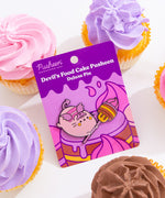Halloween Pusheen Pin attached to backer card. Pin sits on a white surface surrounded by cupcakes with purple, pink, and brown frosting. 