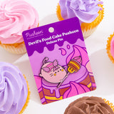 Halloween Pusheen Pin attached to backer card. Pin sits on a white surface surrounded by cupcakes with purple, pink, and brown frosting. 
