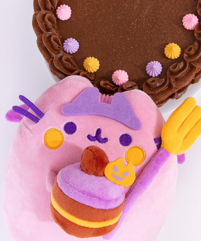 Pusheen Devil's Food Cake Plush lays on a white surface. The light pink and purple plush is holding a fork and cake in her front paws. The crowned plush is surrounded by a chocolate cake decorated with pink, purple, and orange icing dollops. 