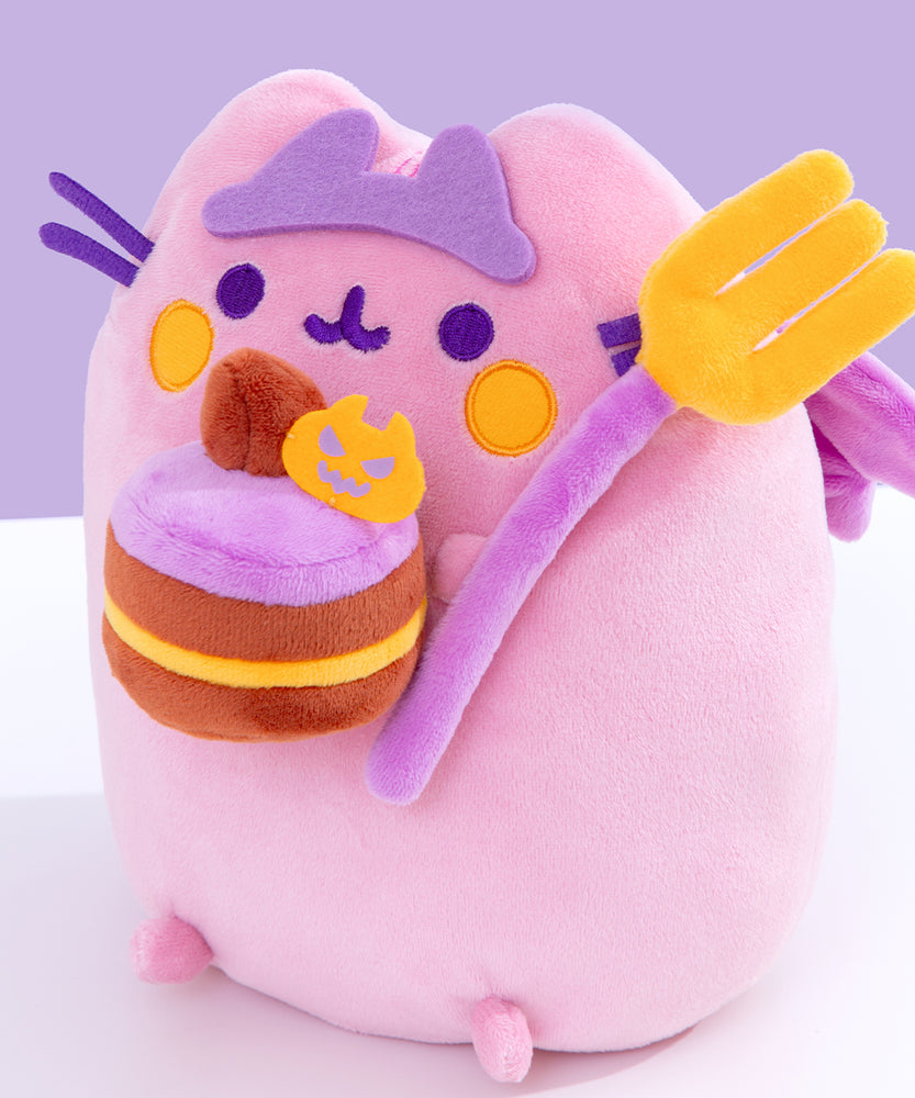 Close-up view of felt crown and the plush items Pusheen is holding. The brown and yellow layered cake plush is topped with purple icing, a chocolate frosting dollop, and a felt orange pumpkin. The large plush fork has a purple handle with yellow tines. 