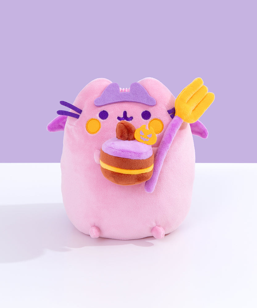 Devil’s Food Cake Pusheen Plush sits atop a white surface in front of a purple background. The pink plush holds a yellow and purple fork and a layered devil’s food cake. 