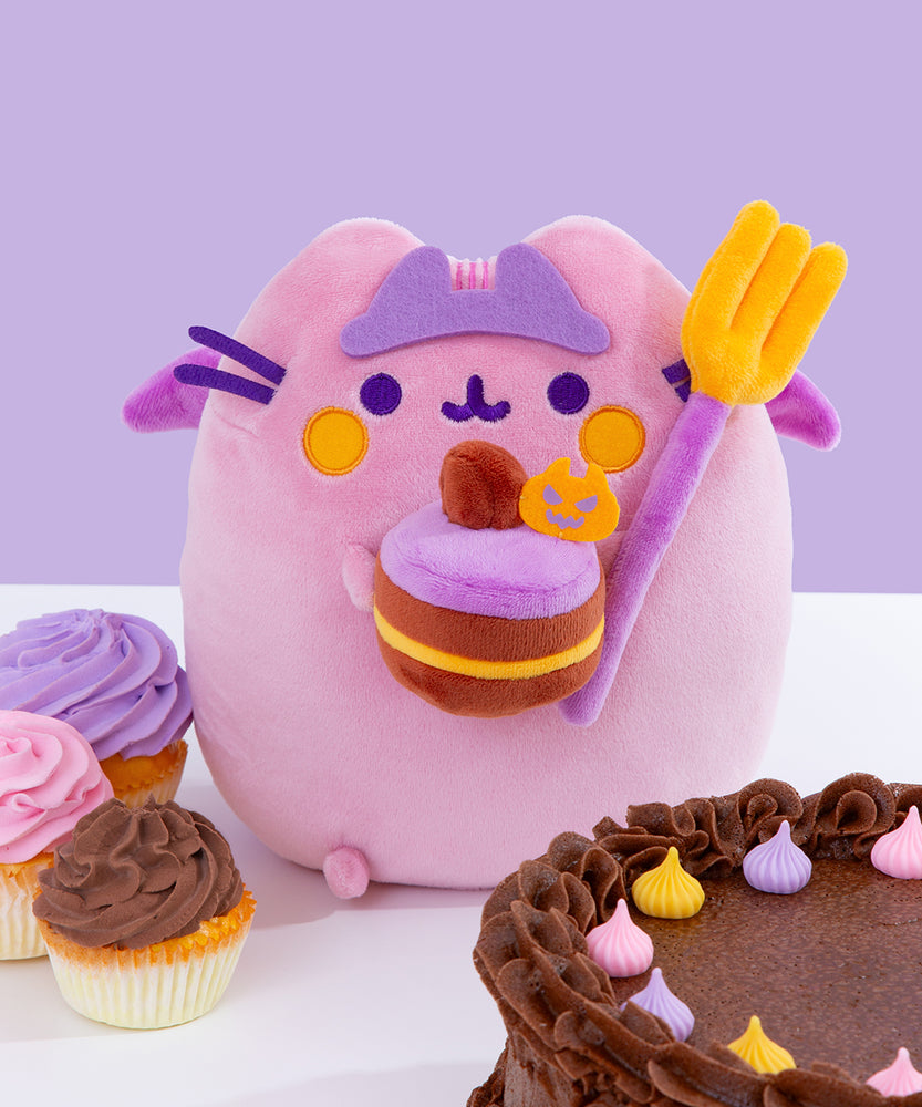 Front view of Devil’s Food Cake Pusheen Plush. The plush a light pink and purple Pusheen smiling. The cat plush wears a light purple crown while holding a purple and yellow fork and a yellow, brown, and purple layered cake. Pusheen’s purple facial features and paws extend off her body. The Plush sits on a white surface surrounded by purple, orange, and pink sweet treats.