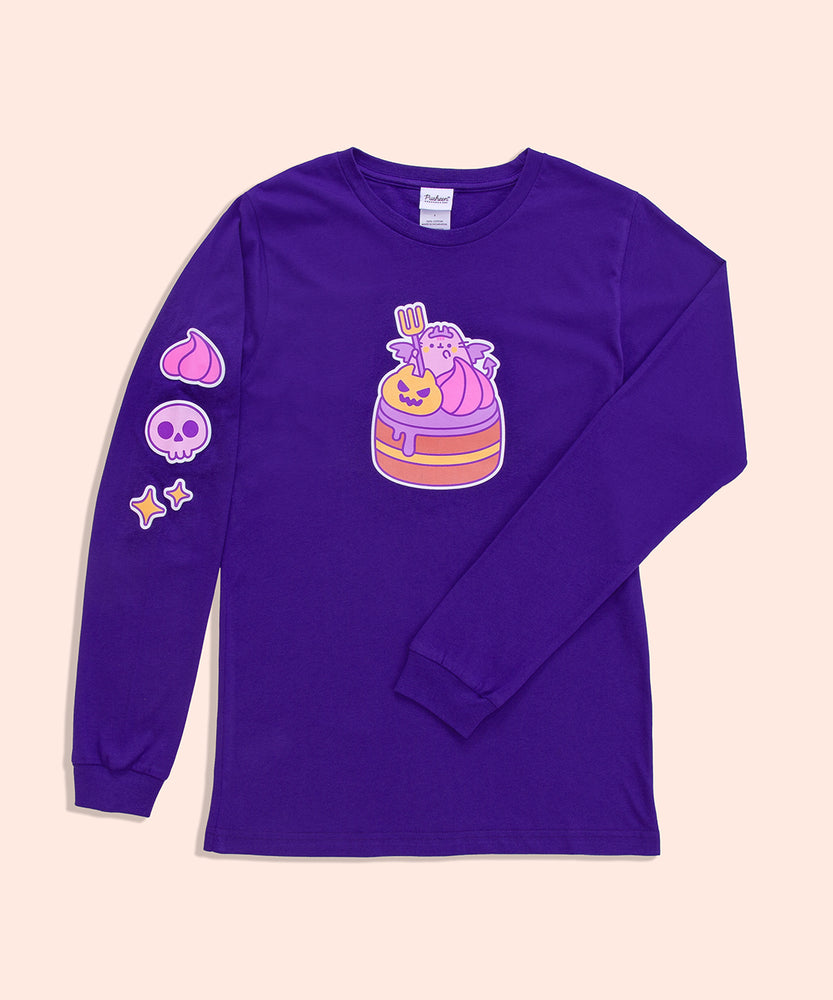 Front view of the Pusheen Devil’s Food Cake Long-Sleeve Tee. The purple graphic t-shirt lays on a light yellow surface. In this view, the large front graphic of Pusheen and her Devil’s Food Cake and the printed graphic detail down the wearer’s right sleeve can be seen. 