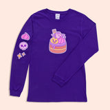 Front view of the Pusheen Devil’s Food Cake Long-Sleeve Tee. The purple graphic t-shirt lays on a light yellow surface. In this view, the large front graphic of Pusheen and her Devil’s Food Cake and the printed graphic detail down the wearer’s right sleeve can be seen. 