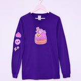 The Devil’s Food Cake Pusheen Long-Sleeve Unisex Tee hangs on a light pink hanger in front of a light purple wall. The dark purple tee shirt features Pusheen the Cat as a flying devil holding a pitch fork while sitting atop a layered devil’s food cake. The large, center front graphic is yellow, pink, purple, orange, and brown is accompanied by a pink whipped dollop, purple skull, and yellow sparkles graphic down the wearer’s right sleeve. 