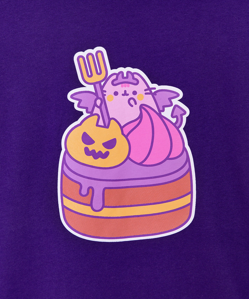 Close-up view of Pusheen devil's food cake graphic tee. A blushing Pusheen holds a fork, wears a crown, and shows off her wings and forked tail while sitting atop a cake behind a scary orange pumpkin and pink frosting dollop. The brown and yellow layered cake has purple frosting dripping down the side. 