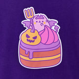 Close-up view of Pusheen devil's food cake graphic tee. A blushing Pusheen holds a fork, wears a crown, and shows off her wings and forked tail while sitting atop a cake behind a scary orange pumpkin and pink frosting dollop. The brown and yellow layered cake has purple frosting dripping down the side. 