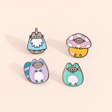 Close-up view of the Enchanted Forest Pins. At the top left is the Gnome pin which showcases Pusheen winking while wearing a gnome costume. At the top right is Pusheen wearing a spotted mushroom costume. At the bottom left is Pusheen as a raccoon with a striped bushy blue and purple tail. At the bottom right is Pusheen dressed as a green, blushing frog. 