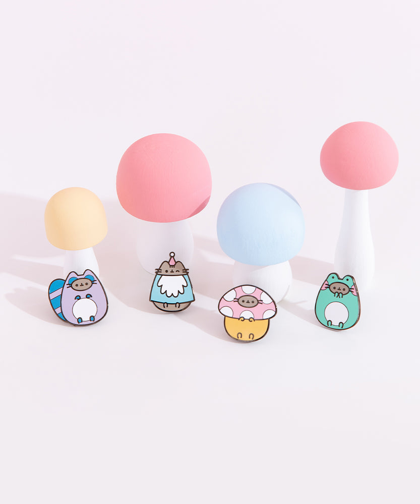 Close-up view of the Enchanted Forest Pins. At the top left is the Gnome pin which showcases Pusheen winking while wearing a gnome costume. At the top right is Pusheen wearing a spotted mushroom costume. At the bottom left is Pusheen as a raccoon with a striped bushy blue and purple tail. At the bottom right is Pusheen dressed as a green, blushing frog. 