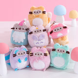 Front view of Pusheen Enchanted Forest Surprise Plush assortment. Pusheen the Cat has taken the form of seven miniature forest creatures in plush form. From left to right and top to bottom, the surprise plush are a Fox, Gnome, Owl, Mushroom, Raccoon, Squirrel and Frog. The keychain plush are surrounded by white, blue, yellow and pink mini mushrooms. 
