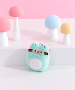 Close-up shot of Pusheen Enchanted Forest character: frog. The plush keychain shows Pusheen the Cat wearing a frog costume. The light green frog is blushing pink and has two eyes that are above Pusheen’s face. 