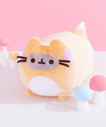 Front view of Pusheen Enchanted Fox Plush. The elongated orange plush is in the shape of a fox leaping. The front of the plush features pink cheek embroidery details with light orange ears extending off the top. On the bottom of the front of the plush is a white embroidery section to mimic a fox’s white stomach. The front of the plush has an oval with Pusheen the Cat’s classic grey and brown features including eyes, mouth, and whiskers. 