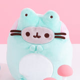 Front view of Pusheen Enchanted Frog Plush. The tall green plush showcases Pusheen the Cat in a frog costume. The front side of the plush features pink cheek embroidery details with light green frog eyes extending off the top. On the bottom of the front of the plush is a white embroidery section to mimic a frog’s white stomach. The front of the plush has an oval with Pusheen the Cat’s classic grey and brown features including eyes, mouth, and whiskers. 