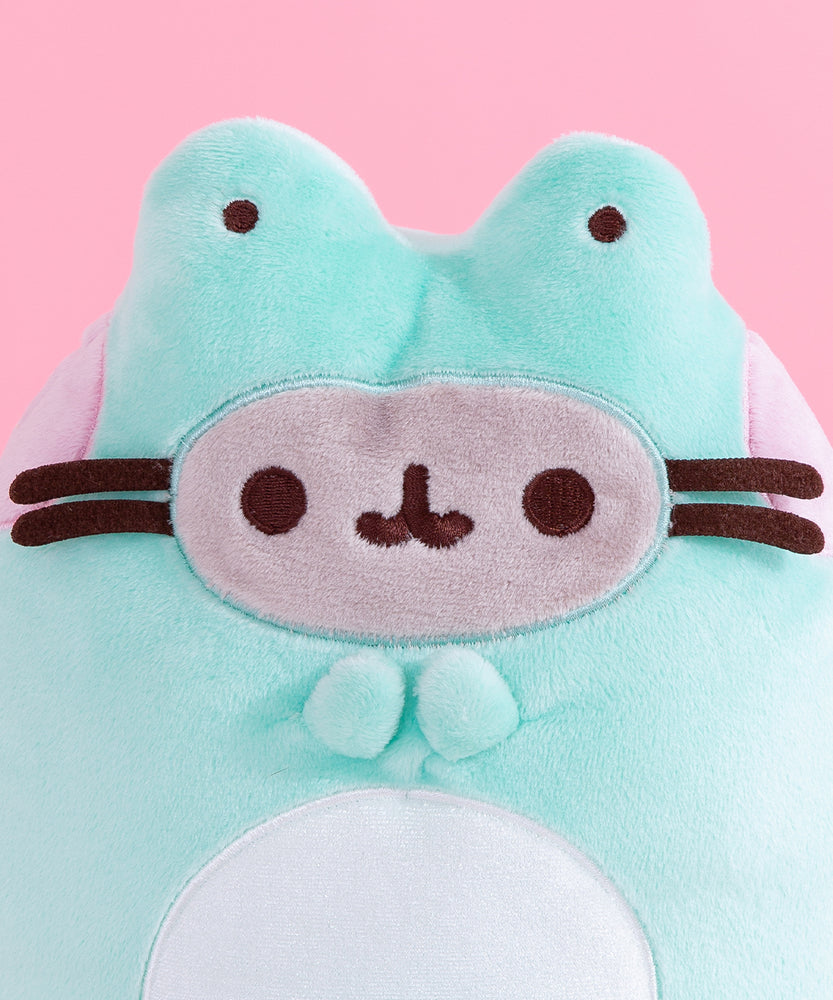 Close-up view of Pusheen Frog plush. Pusheen’s classic grey face and brown facial features are in an oval to mimic that the cat is wearing a frog costume. At the top of the plush are half spheres with brown embroidered eyes to mimic frog’s eyes. 