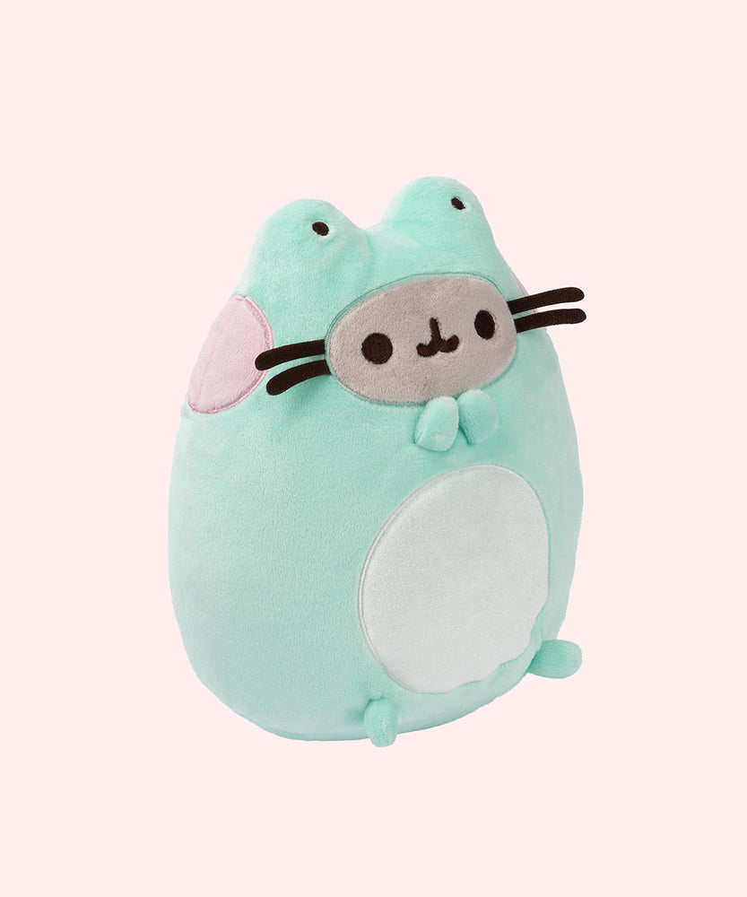 Right side view of the frog Pusheen plush. The green, white, pink, and grey sitting plush is in front of a light pink background. The brown whiskers of the plush extend off of the front of the plush body. 