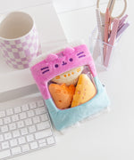 Gamer Pusheen Bag of Chips Plush sits on a white desktop. The pink and blue two-toned bag has a see-through window where the three yellow triangle plush reside.